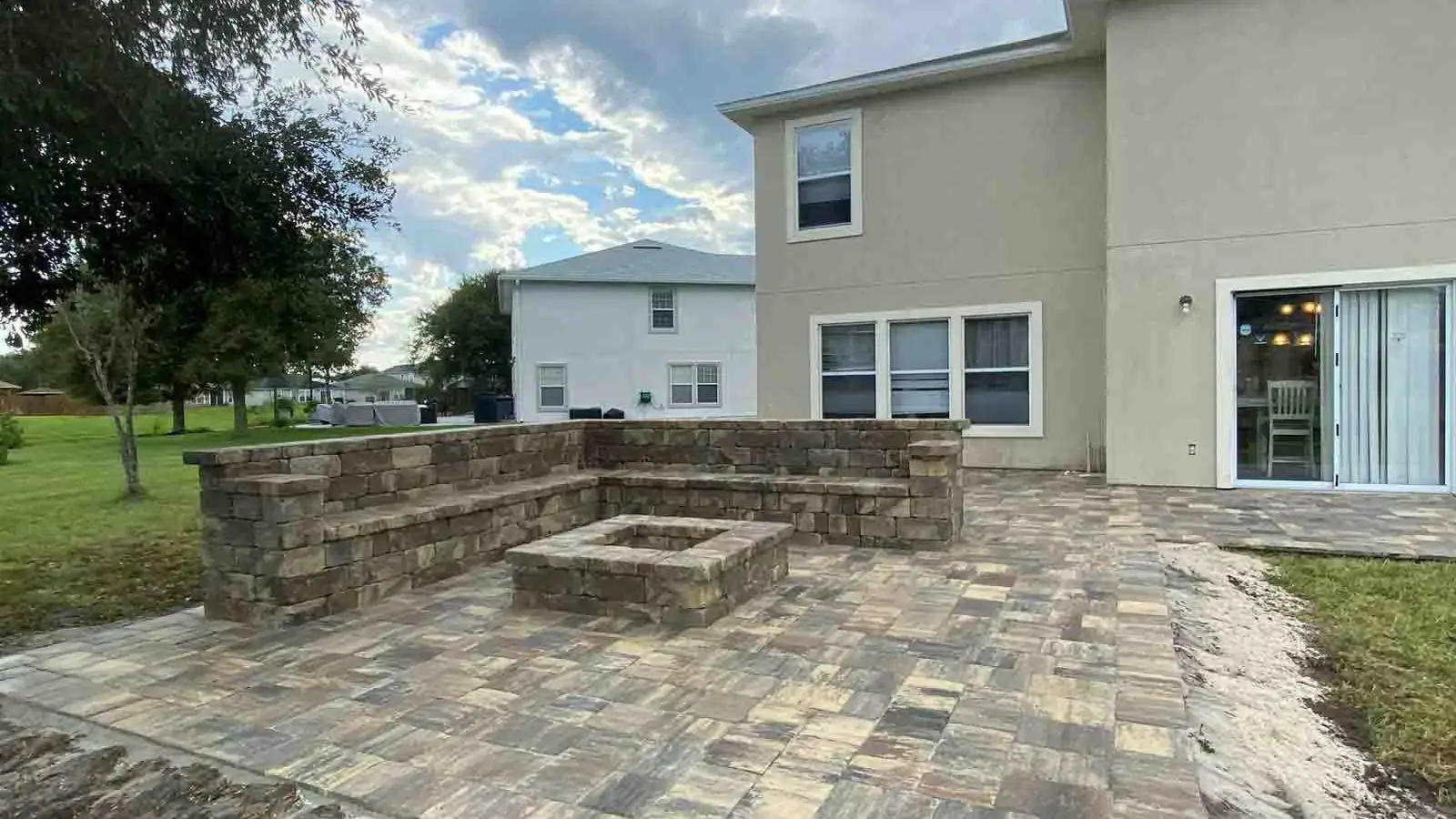 A paver patio with a square fire pit and seating wall behind a two-storey home