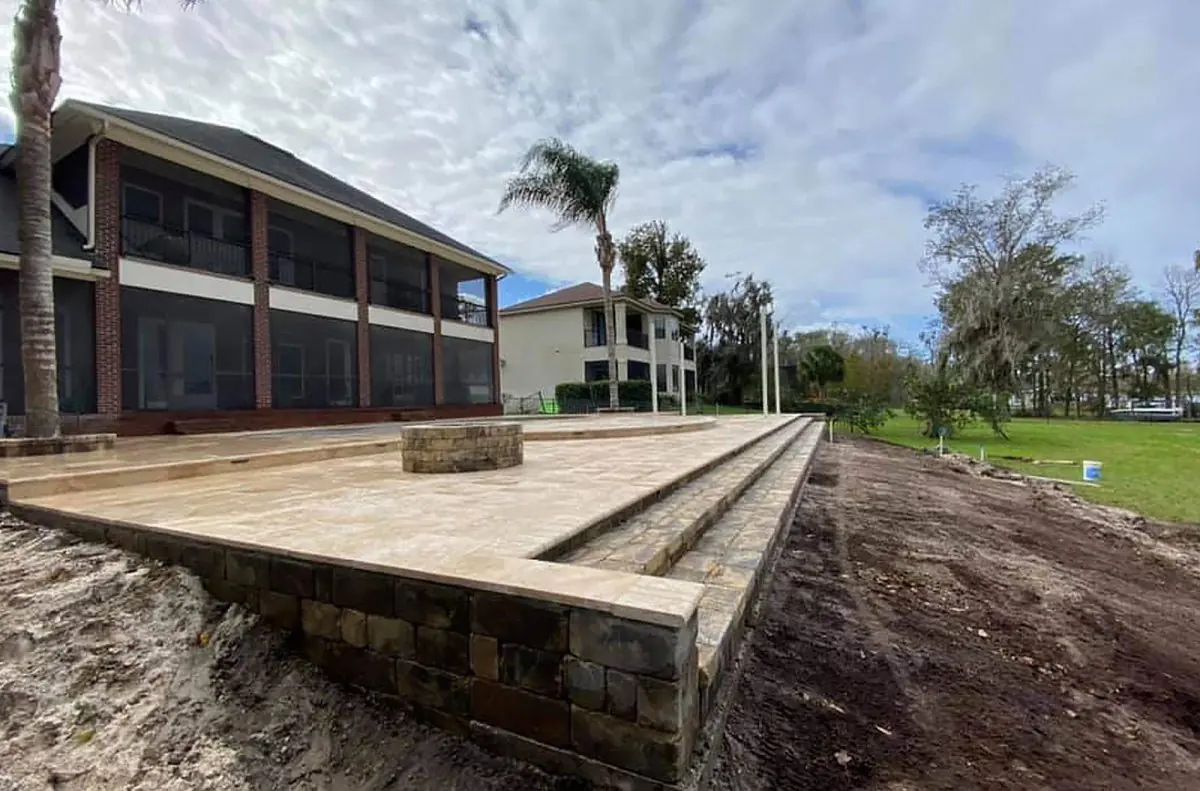 A raised paver patio with steps and a fire pit beside a lake house