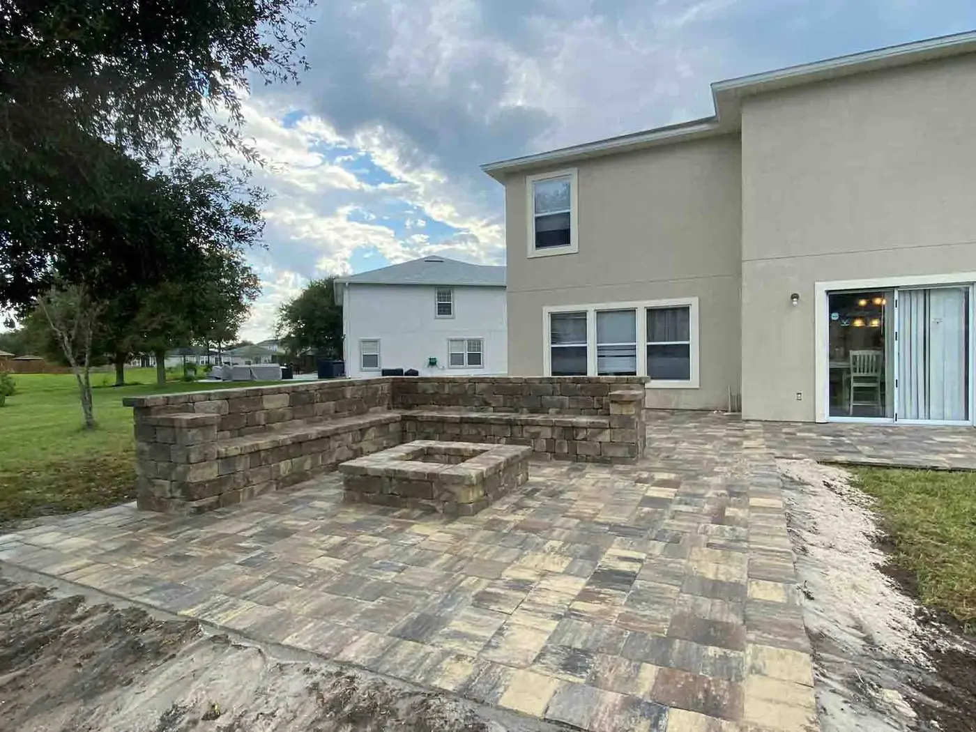 A paver patio with a square fire pit and seating wall behind a two-storey home